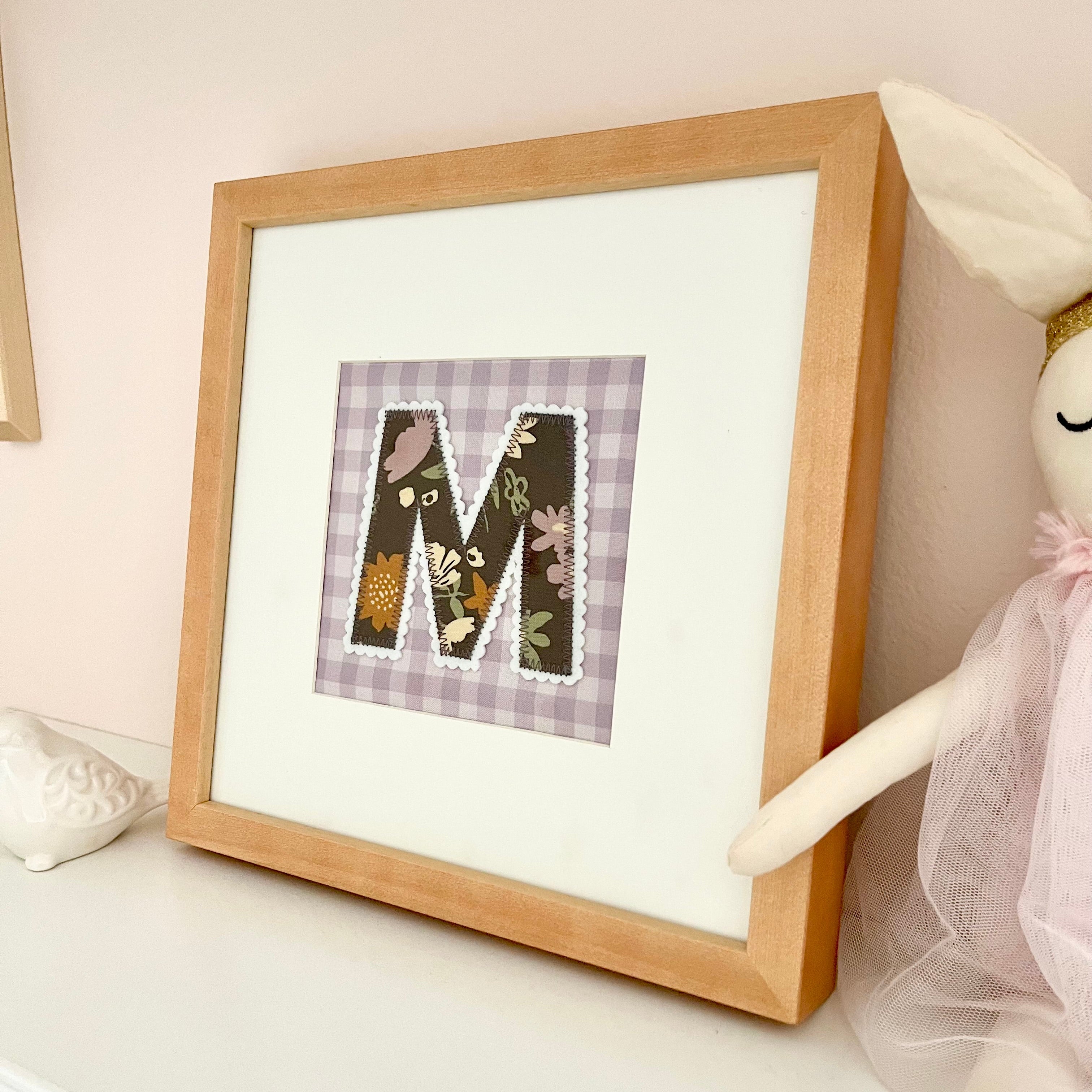 A framed custom monogram letter patchwork art piece with a light wood frame, displayed on a white shelf next to a white bunny doll.